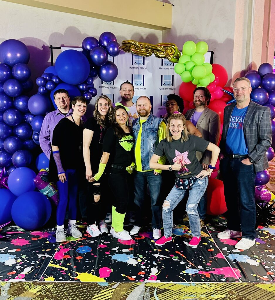 A group of ten people posing together at an event, wearing colorful clothing and accessories. They stand in front of a backdrop decorated with purple and green balloons, with a splattered paint design on the floor.