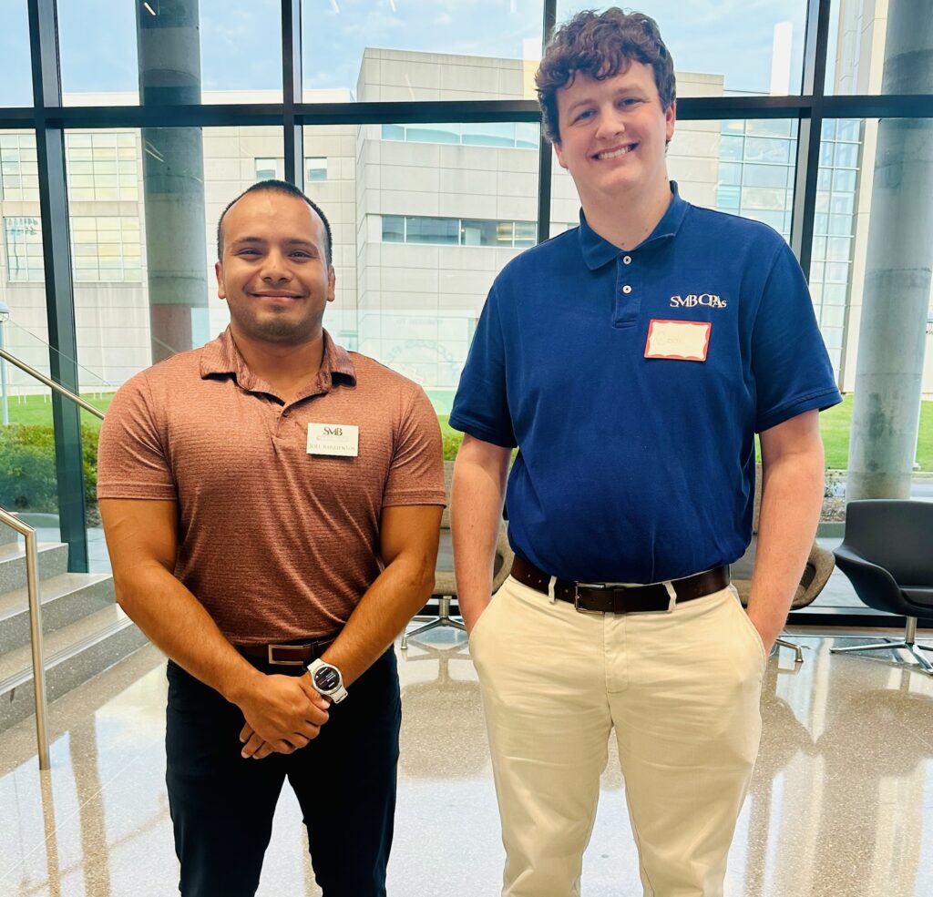 Two men standing side by side in a modern lobby area. The man on the left is wearing a maroon shirt and black pants, while the man on the right is in a blue polo and beige pants. Both are smiling and wearing name tags.