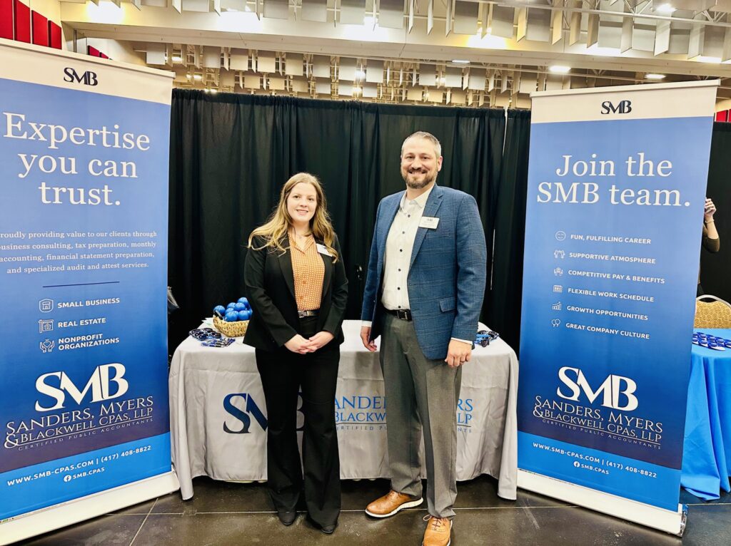 Two professionals standing in front of a booth at a career fair. The booth displays banners promoting Sanders Myers & Blackwell CPAs, highlighting career opportunities. The man is wearing a blue suit and the woman is in a black blazer.