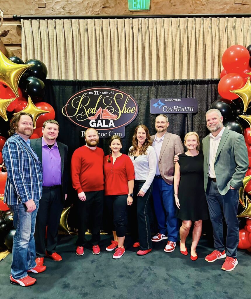 A group of eight people posing for a photo at the 11th Annual Red Shoe Gala. They are wearing red shoes, standing in front of a backdrop with balloons and banners for the event.