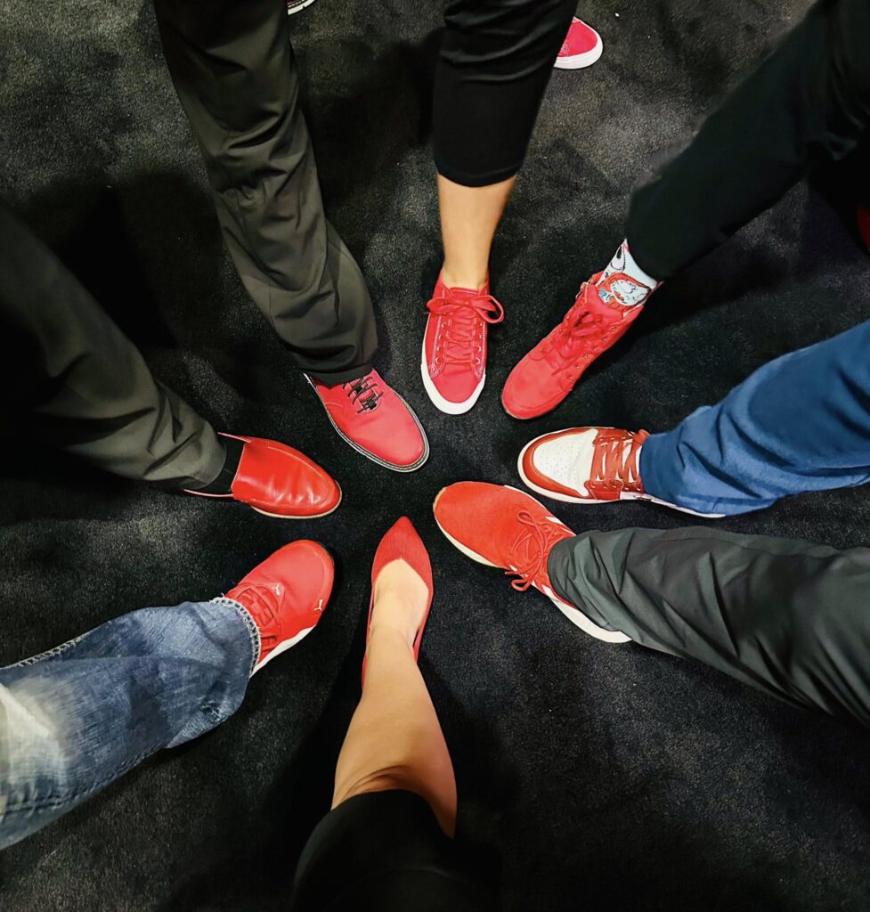 A circle of feet with a variety of footwear on a dark surface. Red sneakers are prominently featured along with a woman's red high-heeled shoe. Some individuals wear jeans and black pants.