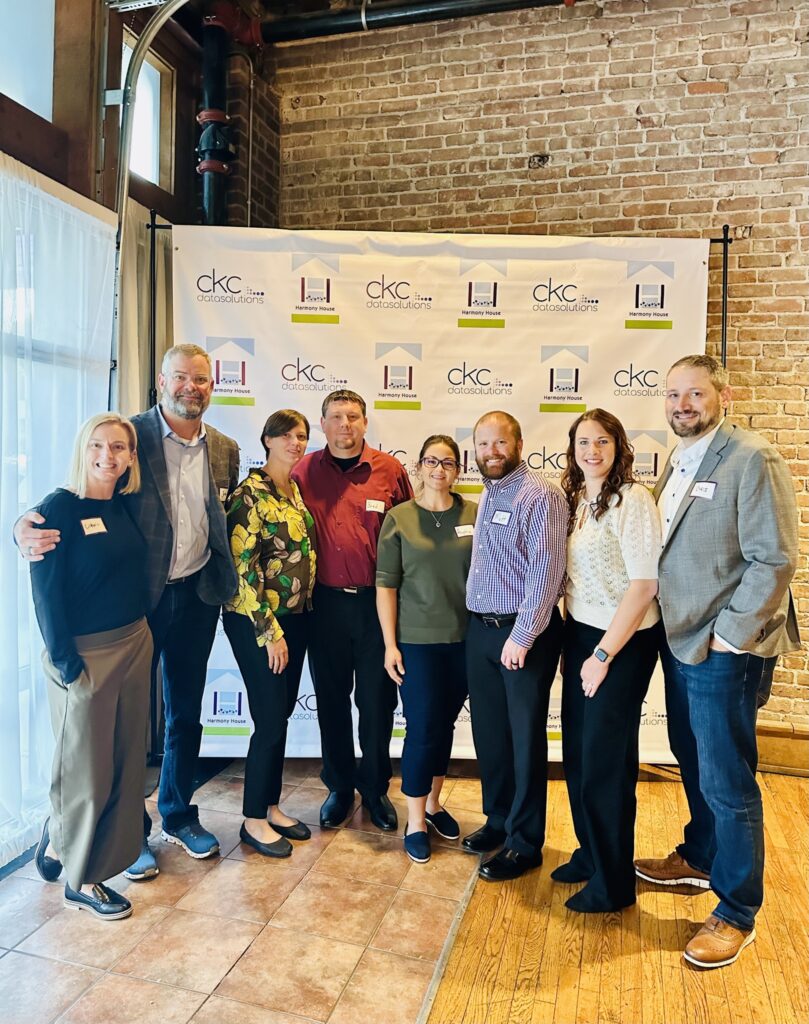 A group of eight individuals poses together in front of a branded backdrop at an event. They are wearing a variety of professional attire and smiling. The backdrop features logos and patterns related to the event.
